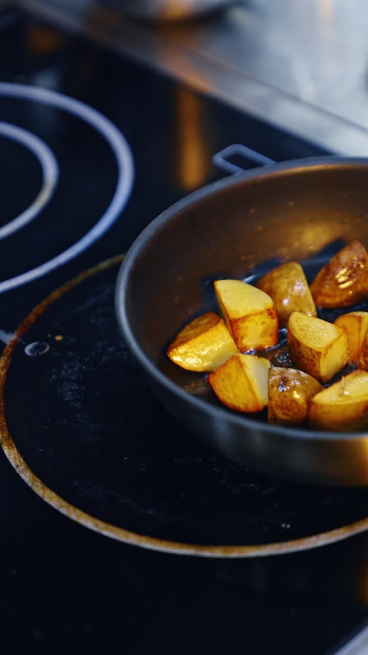 Potatoes cut into cubes are fried golden on the frying pan. Chef cooking the dish moves the frying pan. Close up. Vertical video.