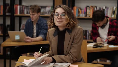 Curly-european-girl-in-brown-jacket-sitting-in-college-library-and-working-on-her-thesis,-preparing-for-exams-then-looks-on-a-camera-and-smiling.-Her-classmates-and-book-shelves-on-the-background