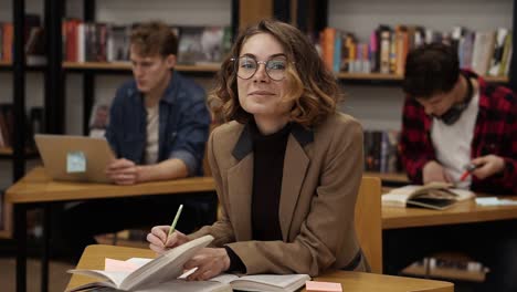 curly european girl in brown jacket sitting in college library and working on her thesis, preparing for exams then looks on a camera and smiling. her classmates and book shelves on the background