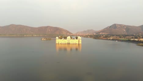 Jal-Mahal-unique-beauty-amidst-the-vast-Sagar-Lake,-reflected-on-the-water-surface-in-Jaipur,-Rajasthan,-India---Aerial-push-in-fly-over-shot