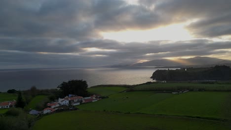 aerial scenic view of sunbeam falling in sea during sunset, drone flying over filed on cliff - azores, portugal