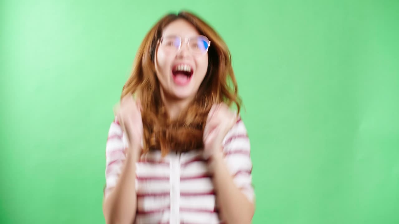 alegre joven asiática con anteojos levantando las manos, temblando y haciendo puños de felicidad, cara de mujer sonriente, fondo de pantalla verde