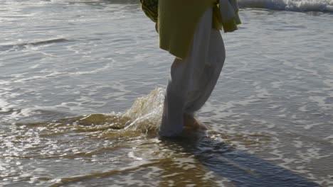 woman with a green sweater tied around her waist and white pants walks along the seashore dragging her bare feet