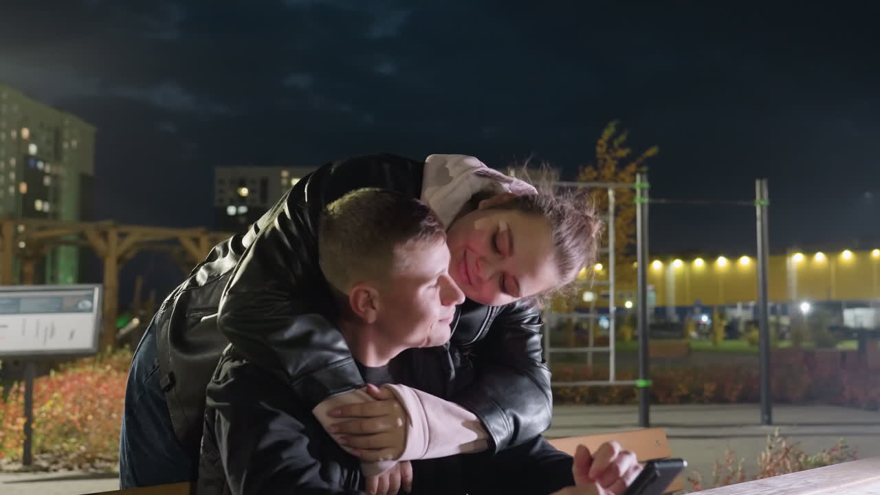 Blogger seated at wooden table operating phone in evening as his sister walks up from behind, gently holds him by neck and shares warm smile with him in peaceful urban park with glowing lights