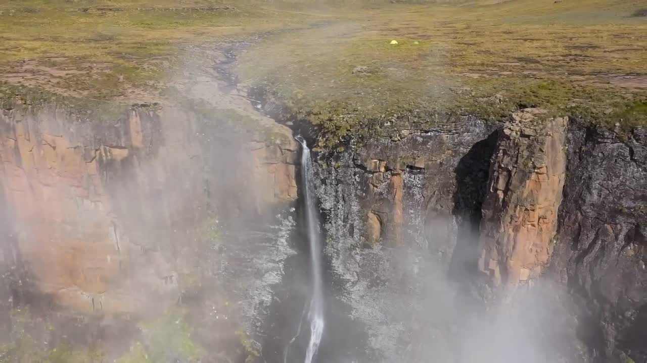 Aerial drone footage flying further backwards from Tugela Falls waterfall during a sunny day in South Africa at Royal National park. Narrow river is falling down a steep tall brown cliff, green nature
