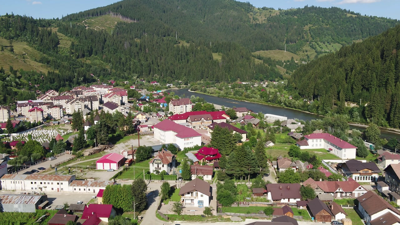 A slow-panning aerial shot reveals a scenic town on a riverbank, nestled deep within a green valley of the Carpathian mountains in Romania. Brosteni, Suceava.