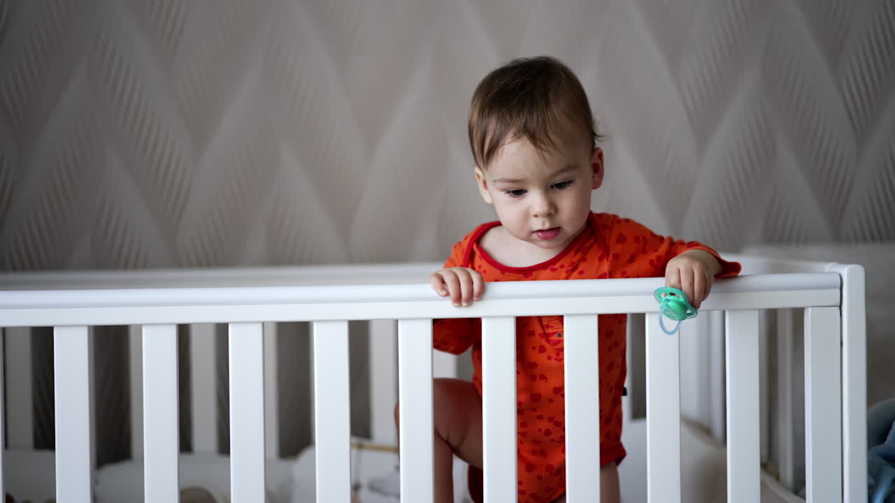 Curious baby boy in orange suit looking down standing in the cot. Little kid starts to smile adorably waving with a pacifier in his hand.