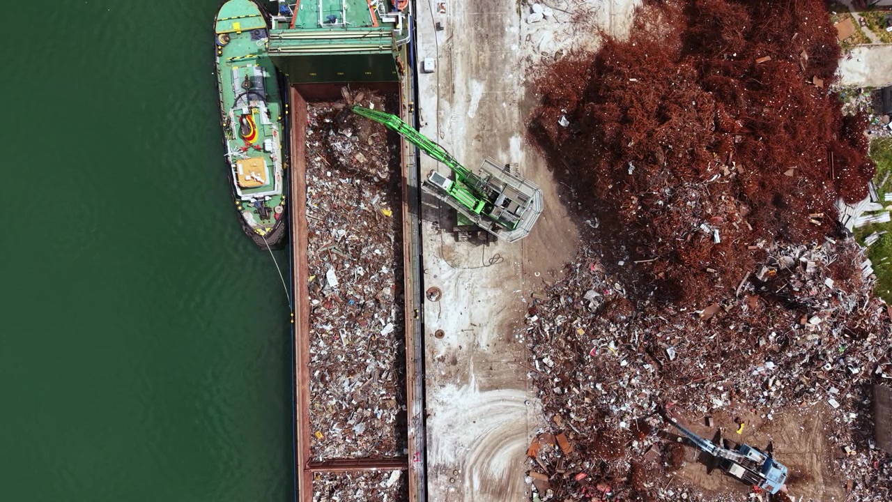Lavori di costruzione in corso presso un impianto di riciclaggio lungo un fiume, con macchinari pesanti che spostano detriti in una giornata di sole.