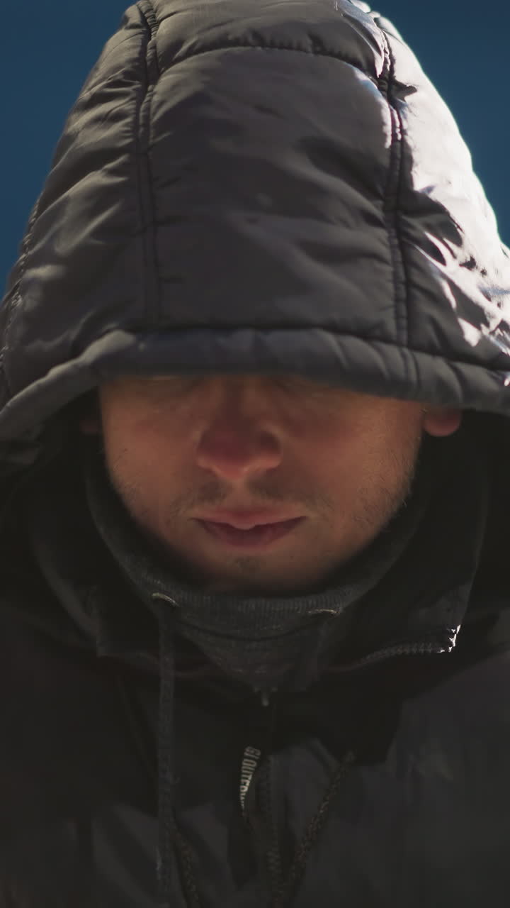 un hombre se sienta al aire libre por la noche en un estadio, apareciendo pensativo, con una capucha cubriendo su cabeza, el fondo presenta una vista borrosa de edificios y luces oscuras