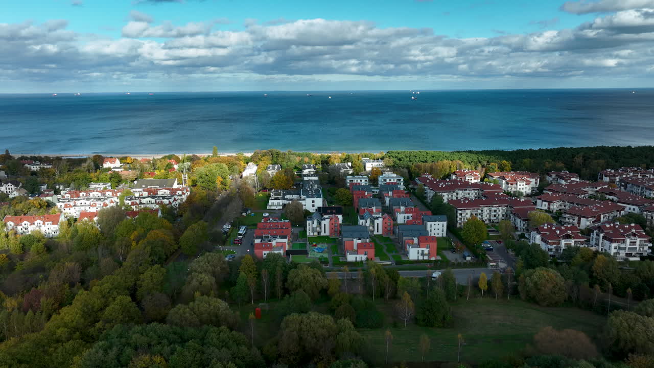 Luftfoto af et kystnært boligområde med moderne bygninger, efterårstræer og Østersøen under en lys, skyet himmel