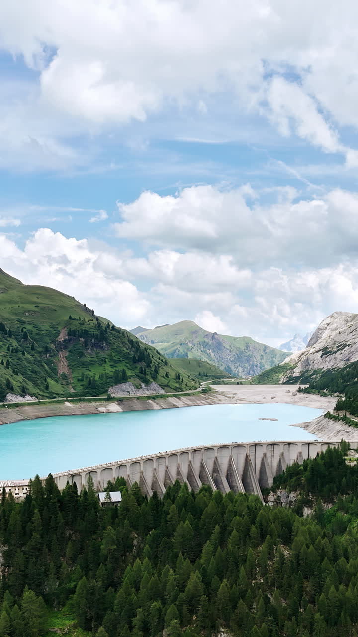 Aerial drone view of a curved concrete dam holding back a turquoise alpine reservoir surrounded by forested mountains and dramatic cliffs in the Italian Alps. Vertical