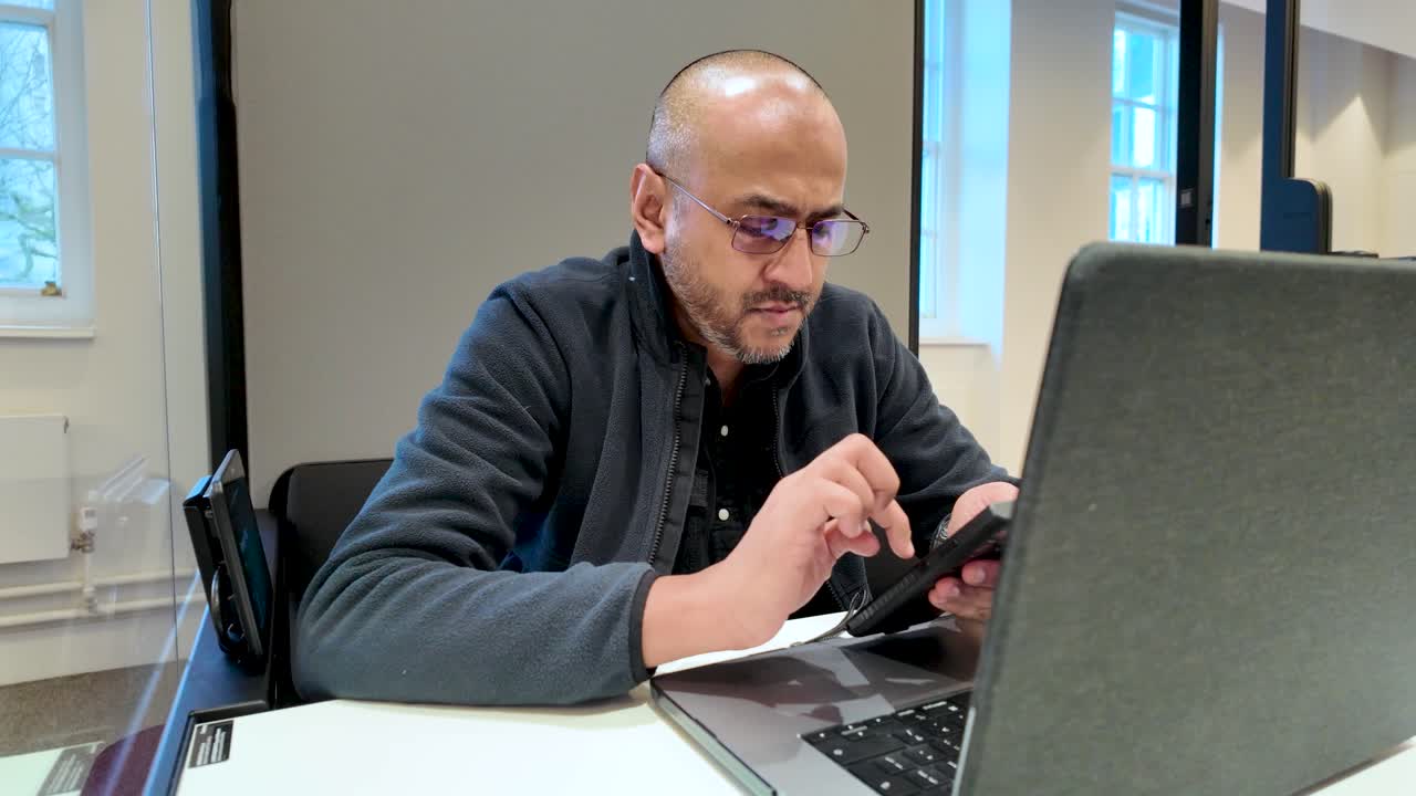 Man using smartphone while seated with laptop inside a contemporary office pod