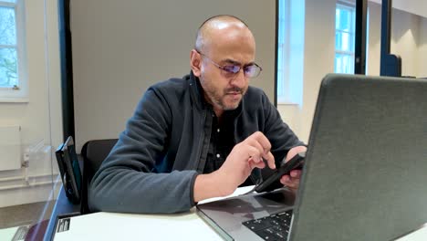 Man using smartphone while seated with laptop inside a contemporary office pod
