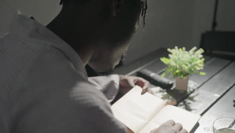 champ reading open book seated at table with potted plant and warm lighting, serene atmosphere. minimalistic indoor space, soft sunlight casting gentle shadows on pages
