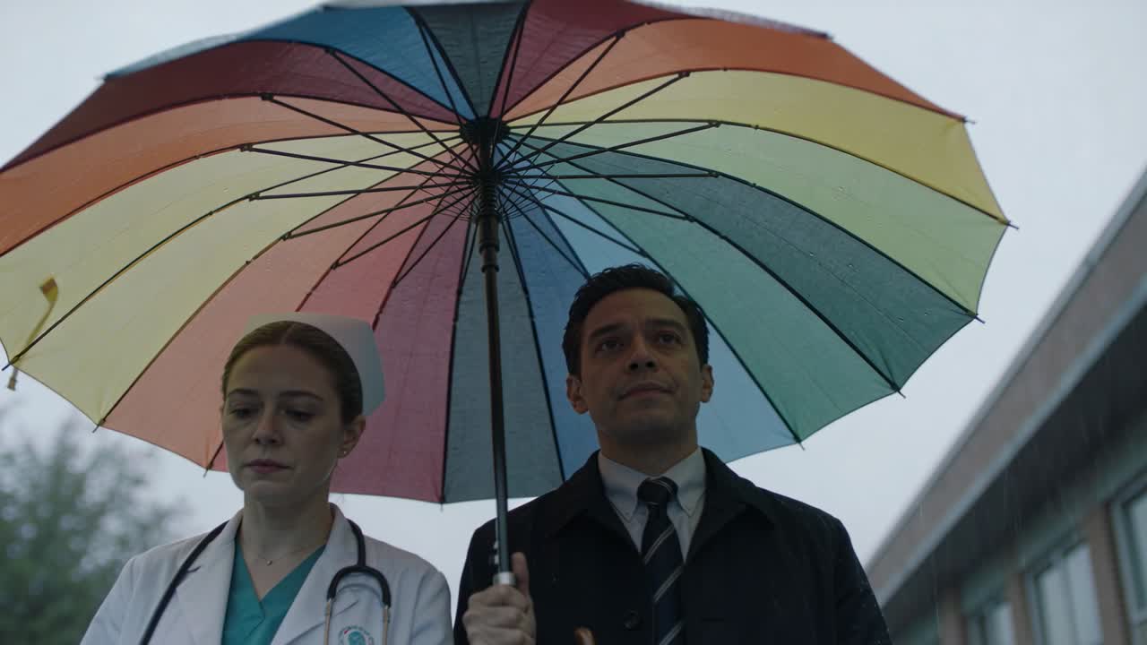 Un uomo cammina tenendo un ombrello arcobaleno, accompagnato da un'infermiera in divisa con uno stetoscopio, all'ospedale mentre cade la pioggia.