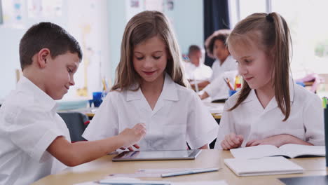 drei grundschüler, die uniform tragen und ein digitales tablet am schreibtisch benutzen