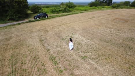 young pregnant woman in a white summer dress standing on a field holding her belly and flowers for a maternity photo shoot - drone helix