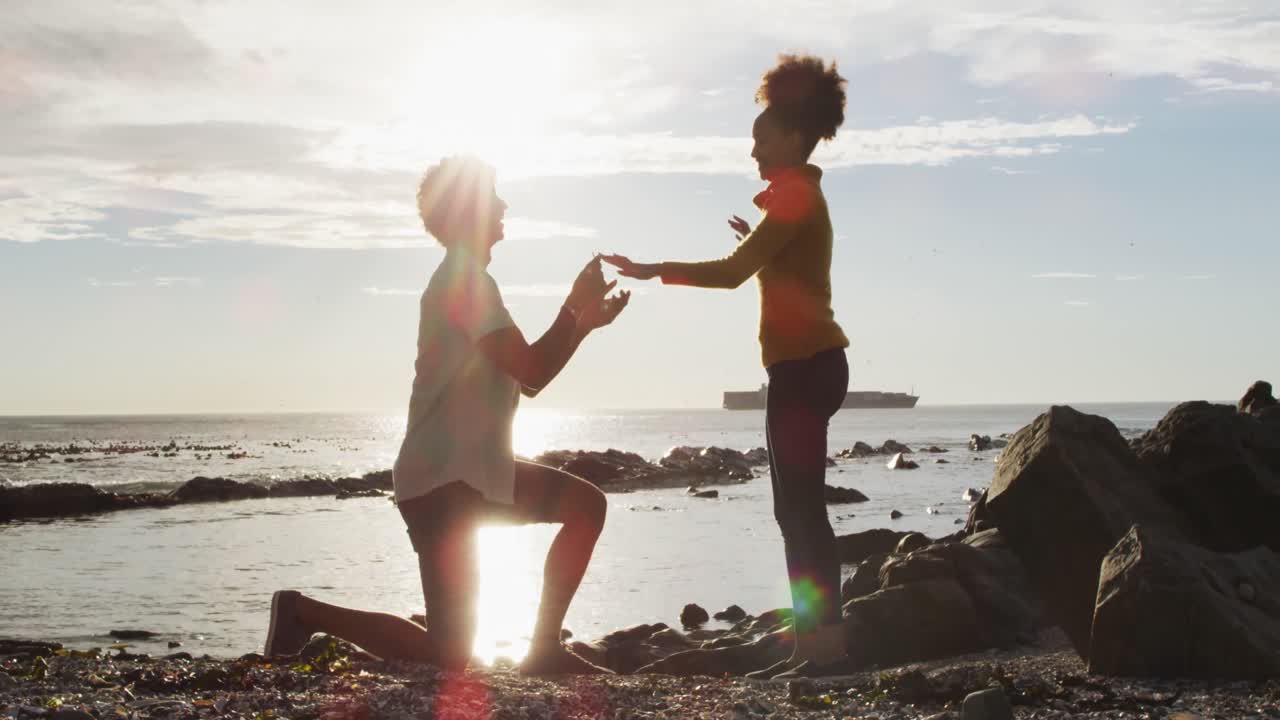 African american man putting a ring on his girlfriend's finger on the rocks near the sea