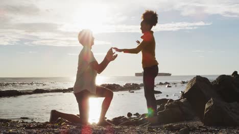 African-american-man-putting-a-ring-on-his-girlfriend's-finger-on-the-rocks-near-the-sea