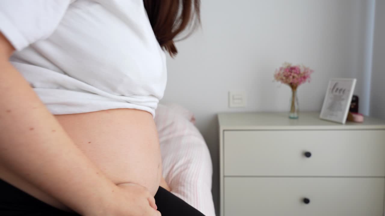 Pregnant woman in bed struggling to get up. Soft light, lactation pillow nearby