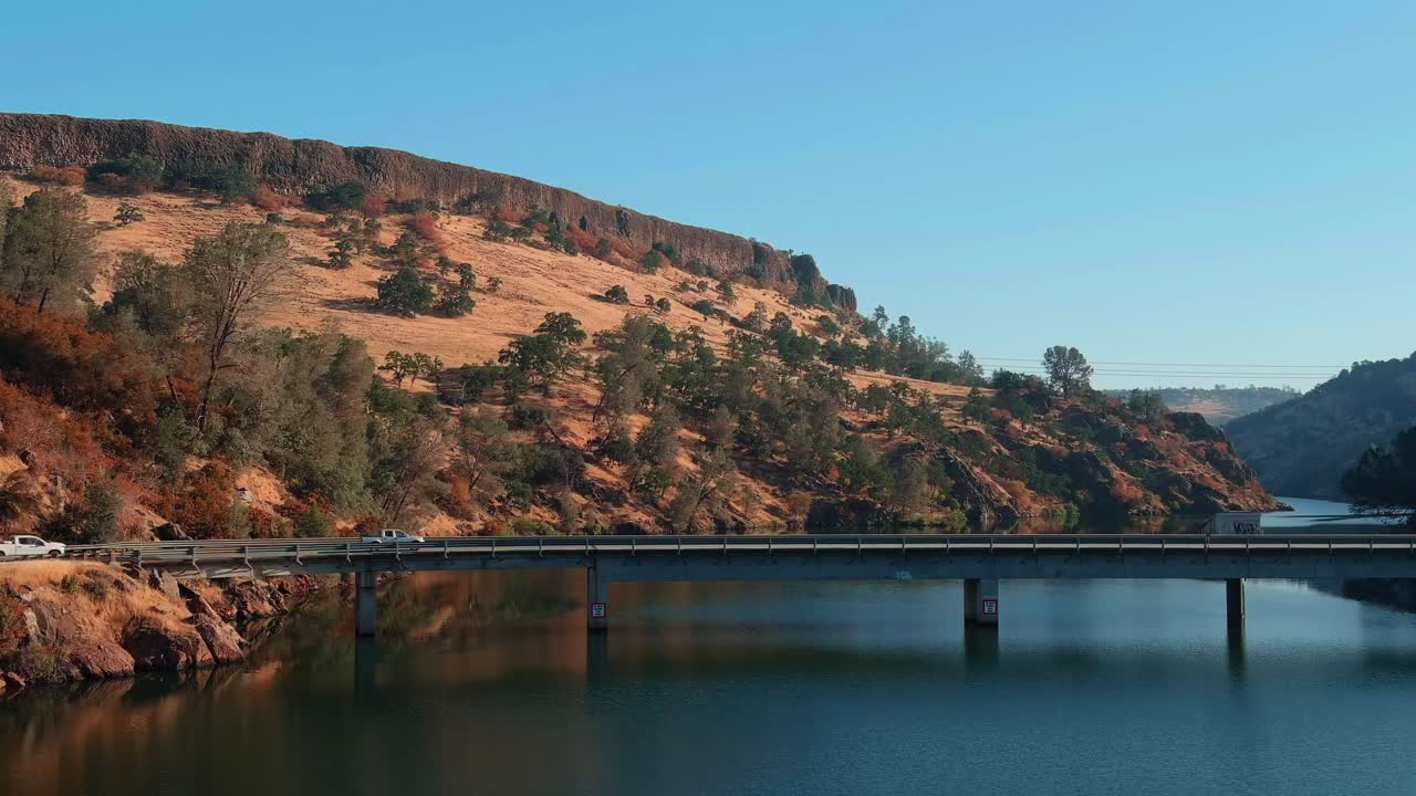 픽업 트럭이 호수를 가로질러 다리 위를 운전하고 있으며, 배경에는 경치 좋은 언덕이 있습니다.