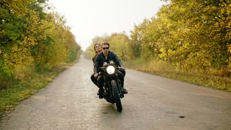 Wide-tracking-shot-of-couple-riding-motorcycle-on-forest-road-in-autumn.-Attractive-young-man-in-sunglasses-driving-his-chopper