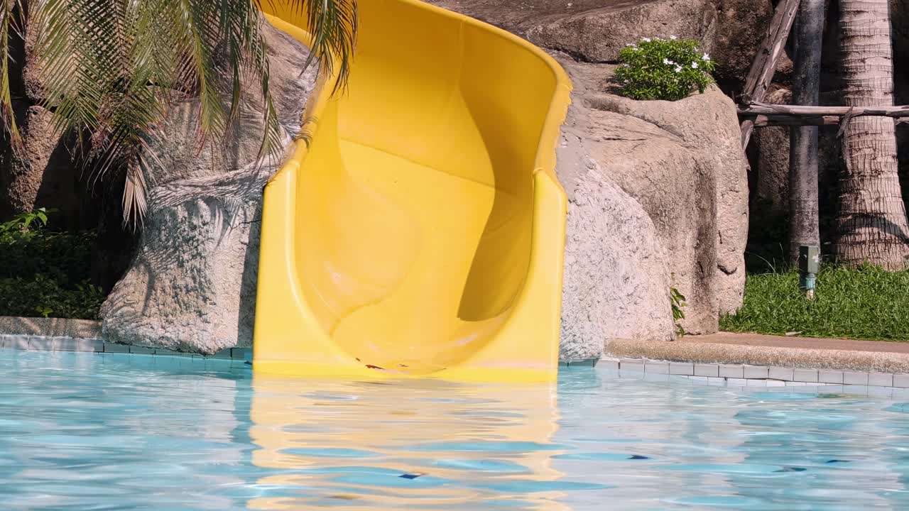 A vibrant yellow slide ends at the edge of a clear blue pool, surrounded by rocks and palm trees.