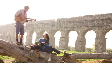 happy young couple backpackers tourists on a log trunk playing guitar singing in front of ancient roman aqueduct ruins in romantic parco degli acquedotti park in rome at sunrise slow motion tripod