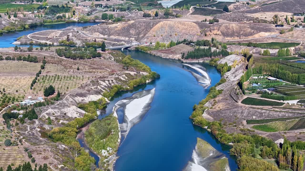 Drone flies high over blue river and bridge with cars, surrounded by fields and hills