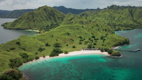 aerial view of komodo island, flores with verdant hills covered in lush greenery. a pristine white sandy beach curves along the bay, with turquoise waters, a wooden pier, and anchored boats.