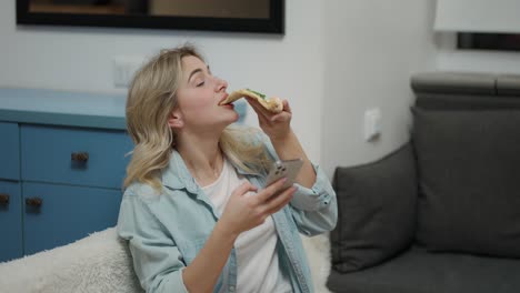 bored girl sitting on sofa at modern living room and taking tasty pizza while use mobile