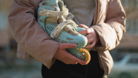 close-up shot of a girl holding a pair of rollerblades with both hands while walking through a sunlit park. she is wearing a peach-colored jacket and the sunlight creates a warm, serene atmosphere