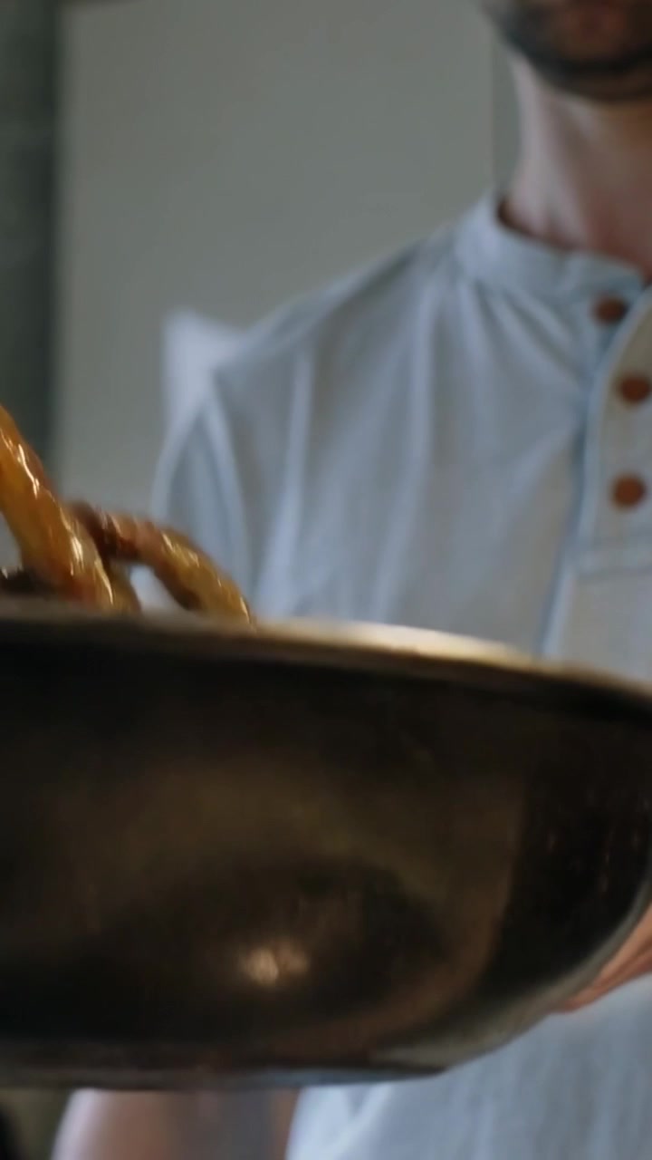 Video verticale: Uomo in camicia blu che regola la presa, solleva e inclina una padella in cucina, girando il cibo.