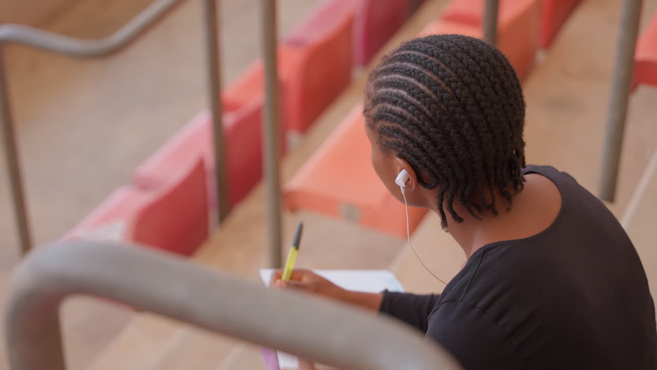 Studente nero che studia sui gradini rossi di uno stadio, bilanciando notebook e telefono, con auricolari che riproducono musica, appoggiato alla ringhiera. Pausa atletica tra gli allenamenti, sguardo concentrato, sfondo di un campus urbano, scatto spontaneo di lato.