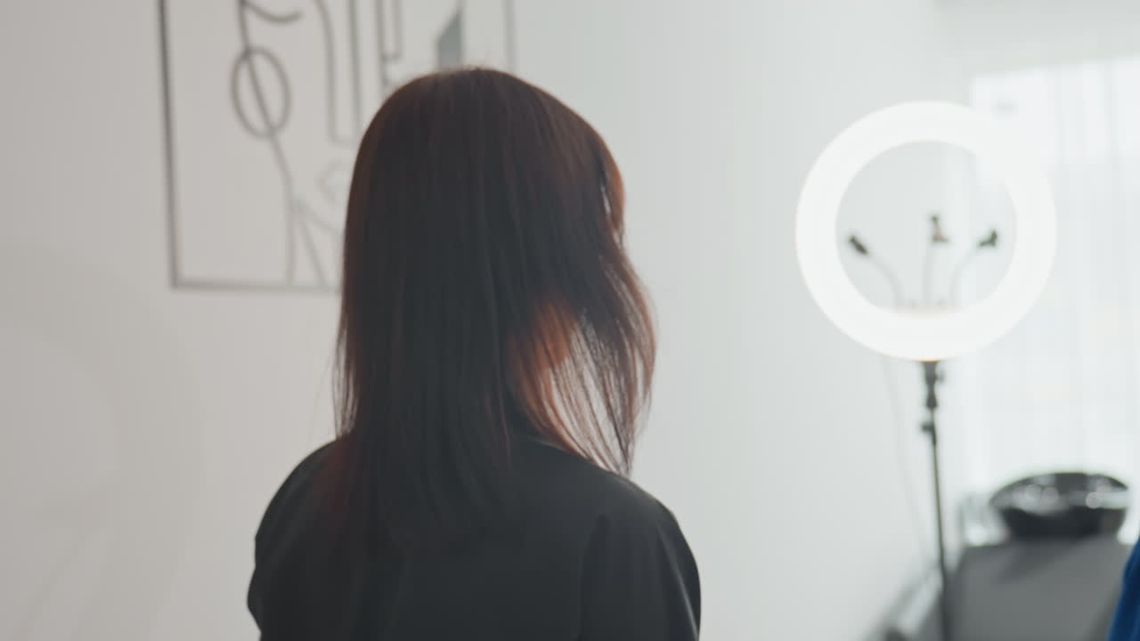 Estilista de peluquería asesorando a un cliente en un estudio luminoso con luz de anillo; el estilista ajusta la capa y habla sobre el corte mientras la luz natural de la ventana ilumina un interior moderno y minimalista, con tonos suaves y un ambiente profesional y tranquilo