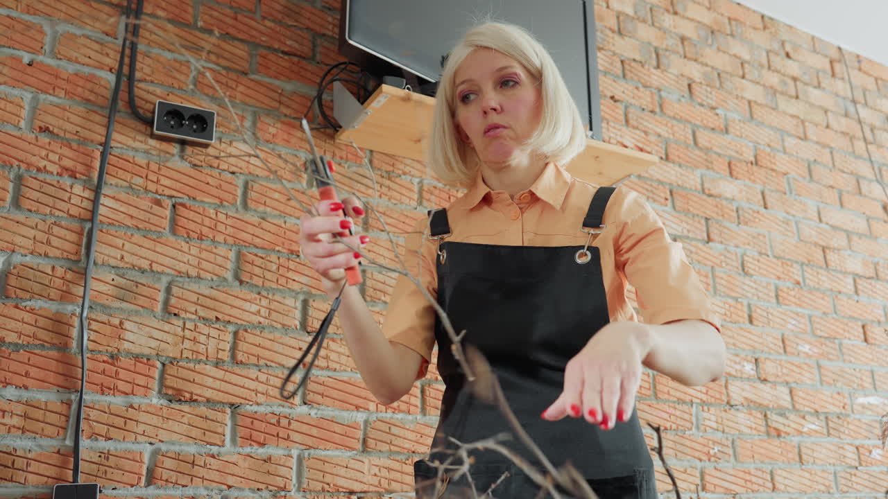 Woman wearing apron holding dry twigs and pruning shears while preparing natural branches for craft project, highlighting creative handmade process with rustic organic materials in artistic indoor