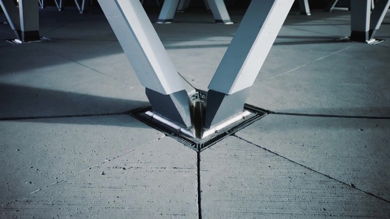 La telecamera si allontana rivelando gambe metalliche a V che si uniscono a una base imbullonata al cemento con drenaggio e ombre.