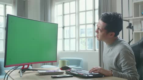 asian man in long sleeved t-shirt with green screen desktop computer having backache while working at home.