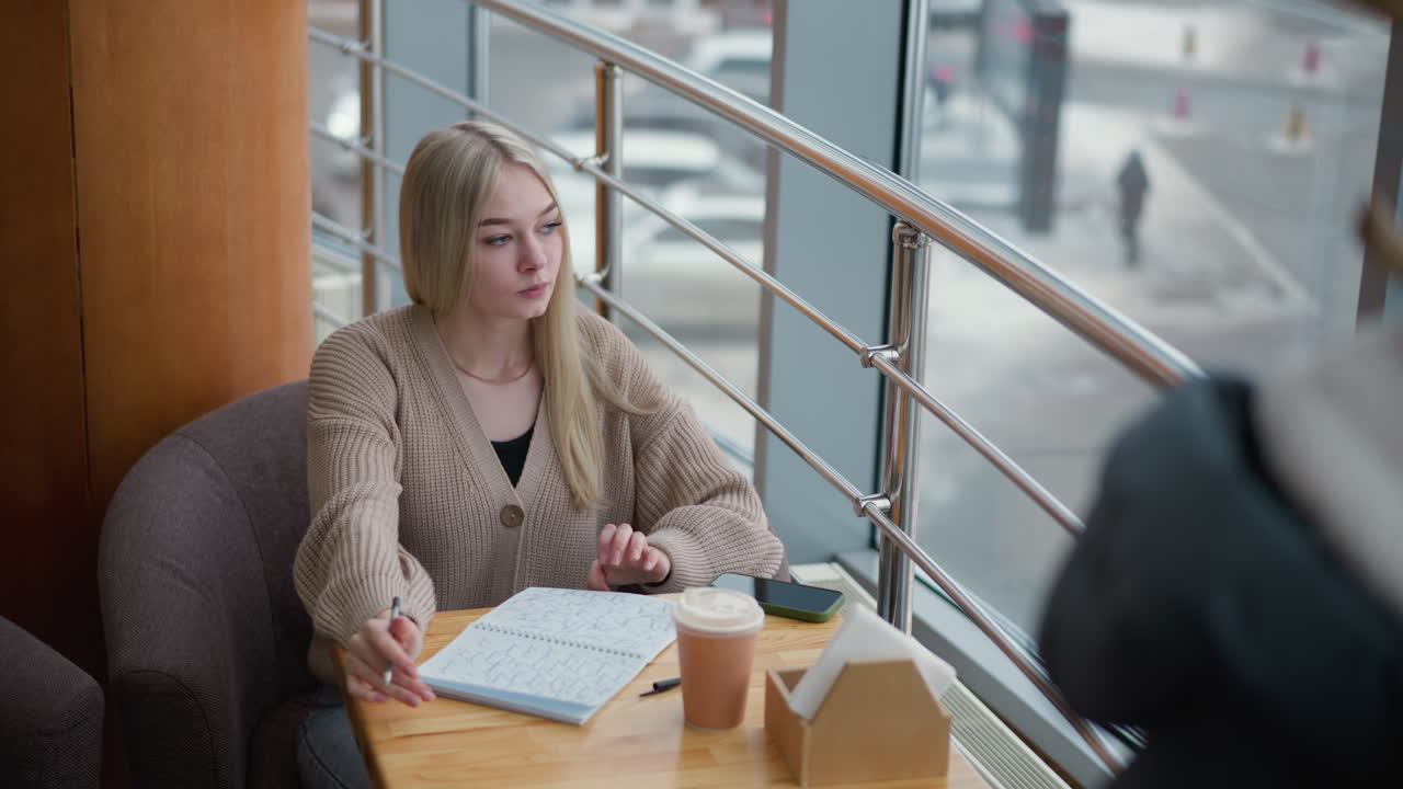 estudiante de secundaria escribiendo pensativo en una cafetería con taza de café en la mesa y ventanas de vidrio que muestran un entorno de invierno, chaqueta de invierno colgada en un estante de tela en el fondo