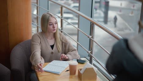 high school student writing thoughtfully in cafe with coffee cup on table and glass windows showcasing a winter setting, winter jacket hanging on cloth rack in background