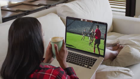 compuesto de una mujer sentada en casa sosteniendo un café viendo un partido de rugby en una computadora portátil