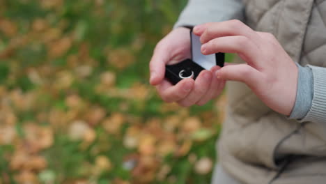 close up of young boy on one knee gently holding ring box with both hands during emotional proposal in outdoor setting with soft blur background featuring swaying green leaves
