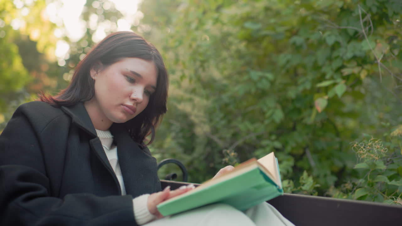 다리를 올리고 공원 벤치에 앉아 초록색 책을 읽고, 페이지 사이에서 머리카락을 정리하고, 땅에는 종이컵이 놓여 있고, 부드러운 가을 햇살이 비추는 가운데 아늑한 코트와 흰색 바지를 입고 조용한 야외에서 휴식을 취하는 차분한 젊은 여성