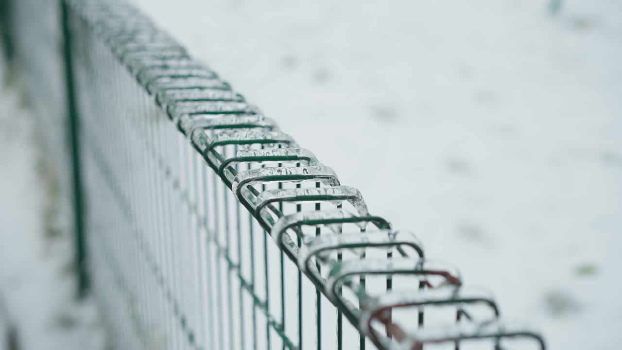 Close-up of a green chain-link fence covered in icicles, set against a blurred snowy landscape, emphasizing the stark coldness of winter with intricate icy textures