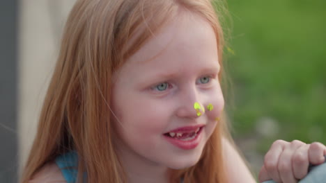 close up ginger kid with neon paint dots on nose and cheek smiling widely while holding paint brush during playful outdoor art session by riverside park in warm golden evening light