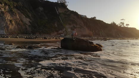 A-man-at-sunrise-watching-the-epic-ocean-waves-crash-on-the-beach-on-the-coast-of-Santa-Barbara,-California-AERIAL-DRONE