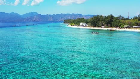 Calm-clear-water-of-turquoise-lagoon-over-rocky-sea-bottom,-boats-floating-near-shore-of-tropical-island-on-a-bright-sky-background