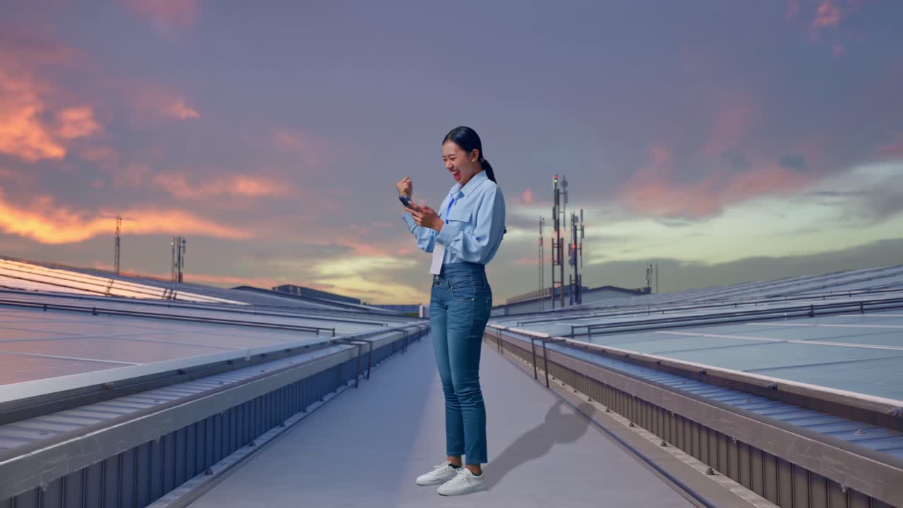 Vista laterale a figura intera di una donna asiatica con il suo telefono, che ispeziona pannelli solari industriali sul tetto, alza il pugno e grida 'gol'.