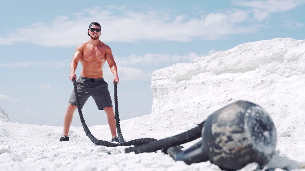 A strong muscular athlete with glasses makes strength exercises for the body with a rope. Shirtless bodybuilder training
