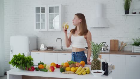 Girl eating banana indoors. Young woman on the modern kitchen background. Healthy fruit and vegetables on the table. Attractive female vegetarian is eating fruit.